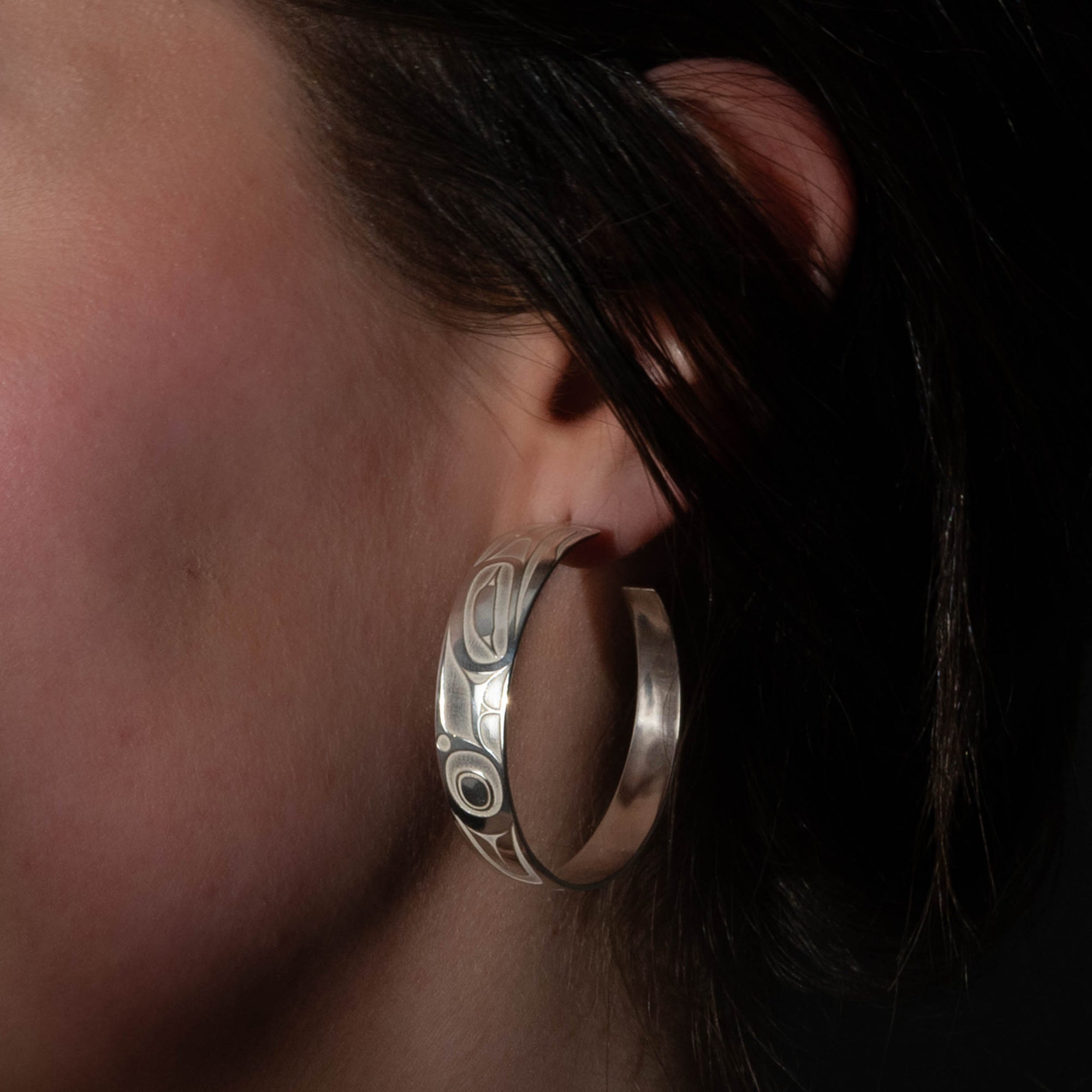 Pair of silver hoop earrings with intricate formline Ts'ymsen thunderbird designs worn by model close up