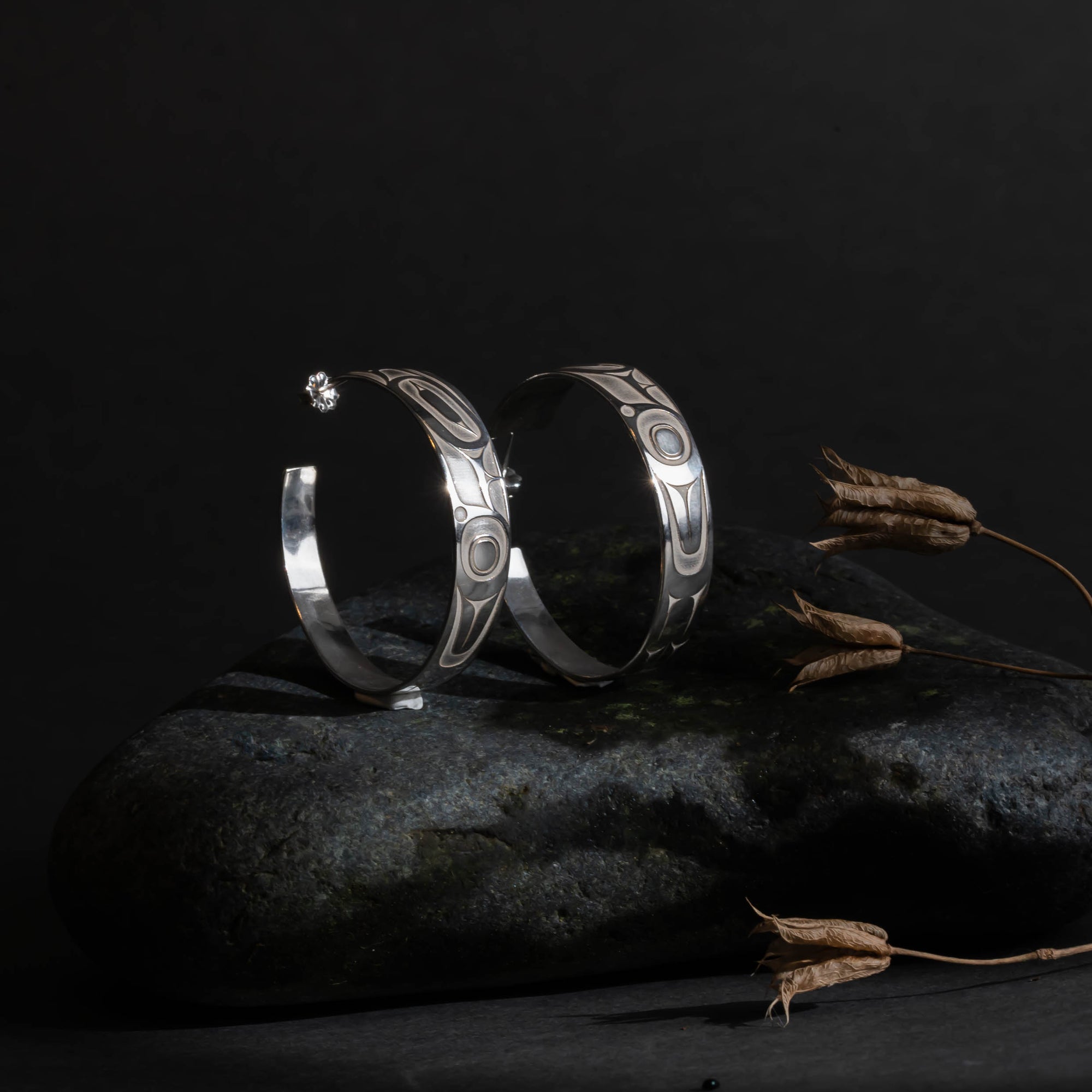 Pair of silver hoop earrings with intricate formline Ts'ymsen thunderbird designs in a room set up