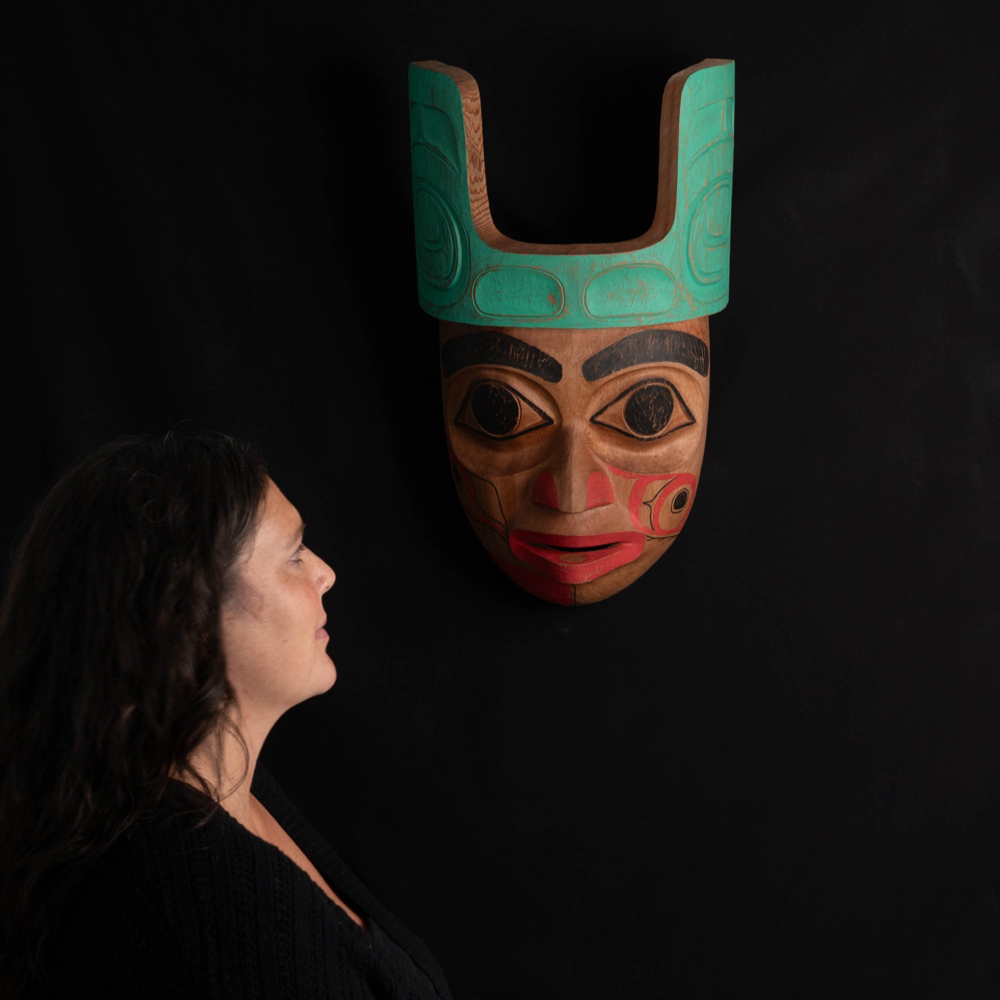 Woman looking at a traditional Ornate Cedar Mouse Woman mask with teal top and black and red face done by Haida artist Corey Bulpitt on a black background