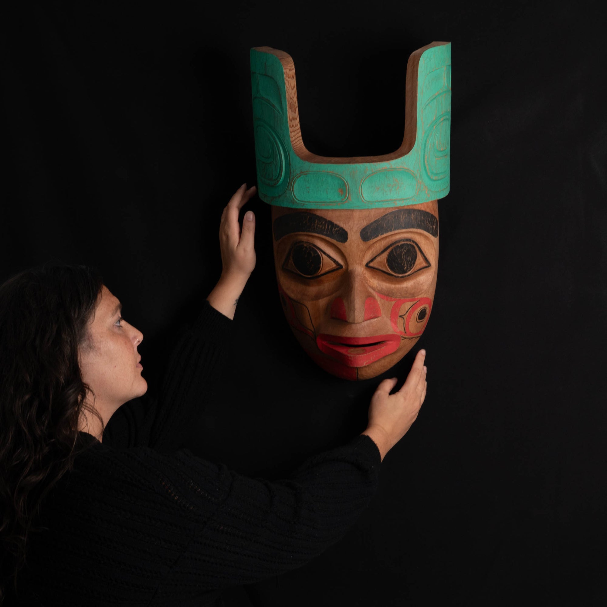 Person holding a large Ornate Cedar Mouse Woman mask with teal top and black and red face done by Haida artist Corey Bulpitt on a black background