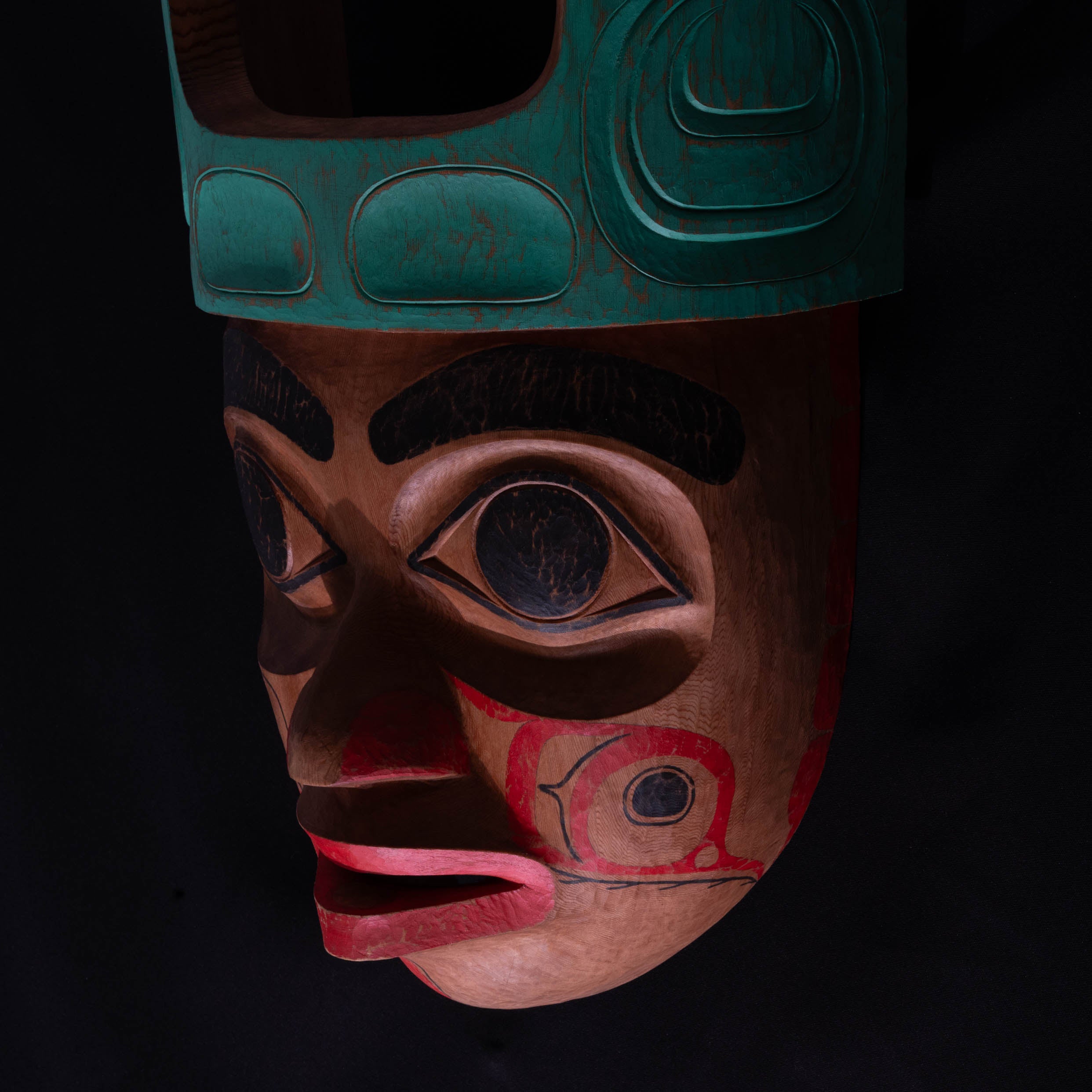 Close up of Cedar Mouse Woman mask with teal top and red face painted and carved by Corey Bulpitt on a black background