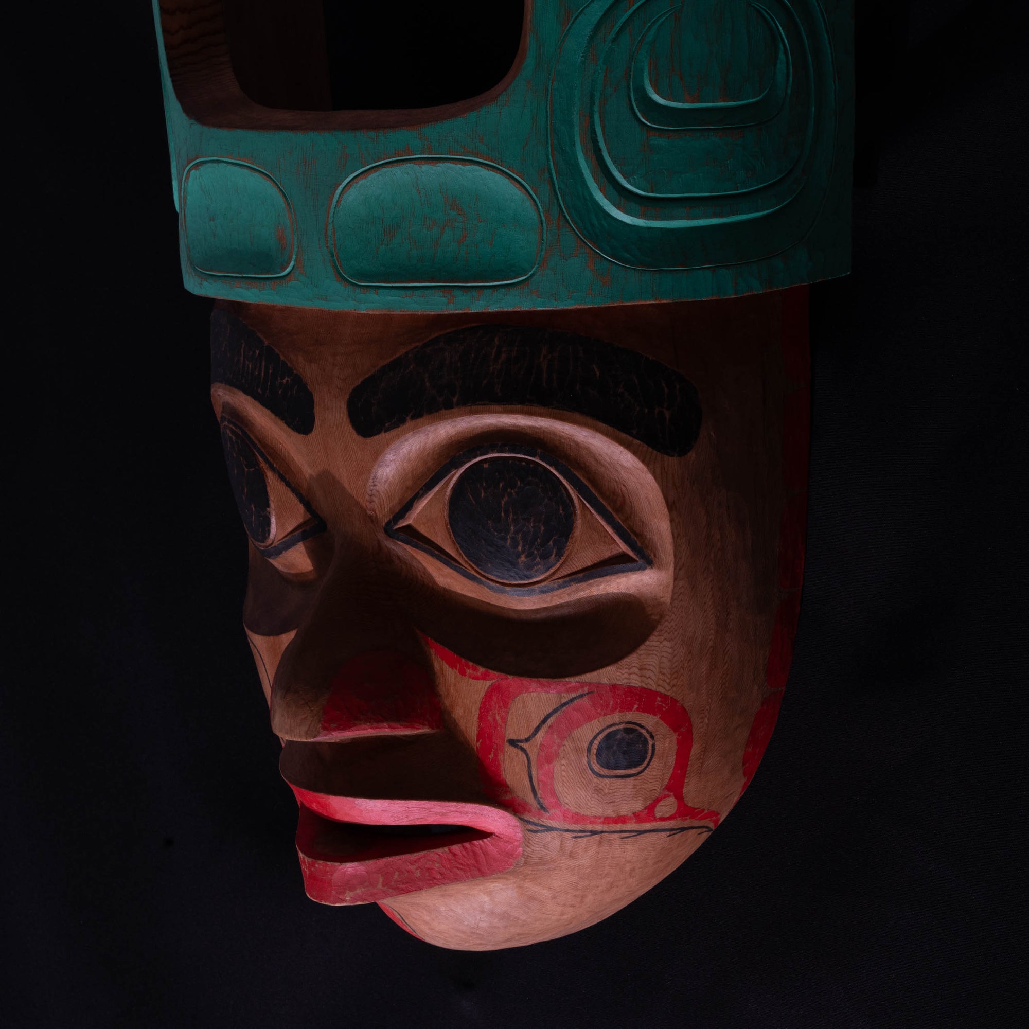 Close up of Cedar Mouse Woman mask with teal top and red face painted and carved by Corey Bulpitt on a black background