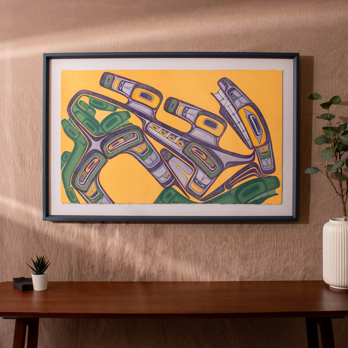 Framed abstract artwork with green and purple design on a yellow background, displayed on a wall above a wooden table.