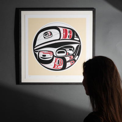 Framed artwork with a stylized Haida Formline Raven design on a beige background, viewed by a person.