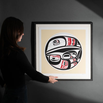 Person touching a framed artwork of a stylized Haida formline Raven with red and black details on a beige background.