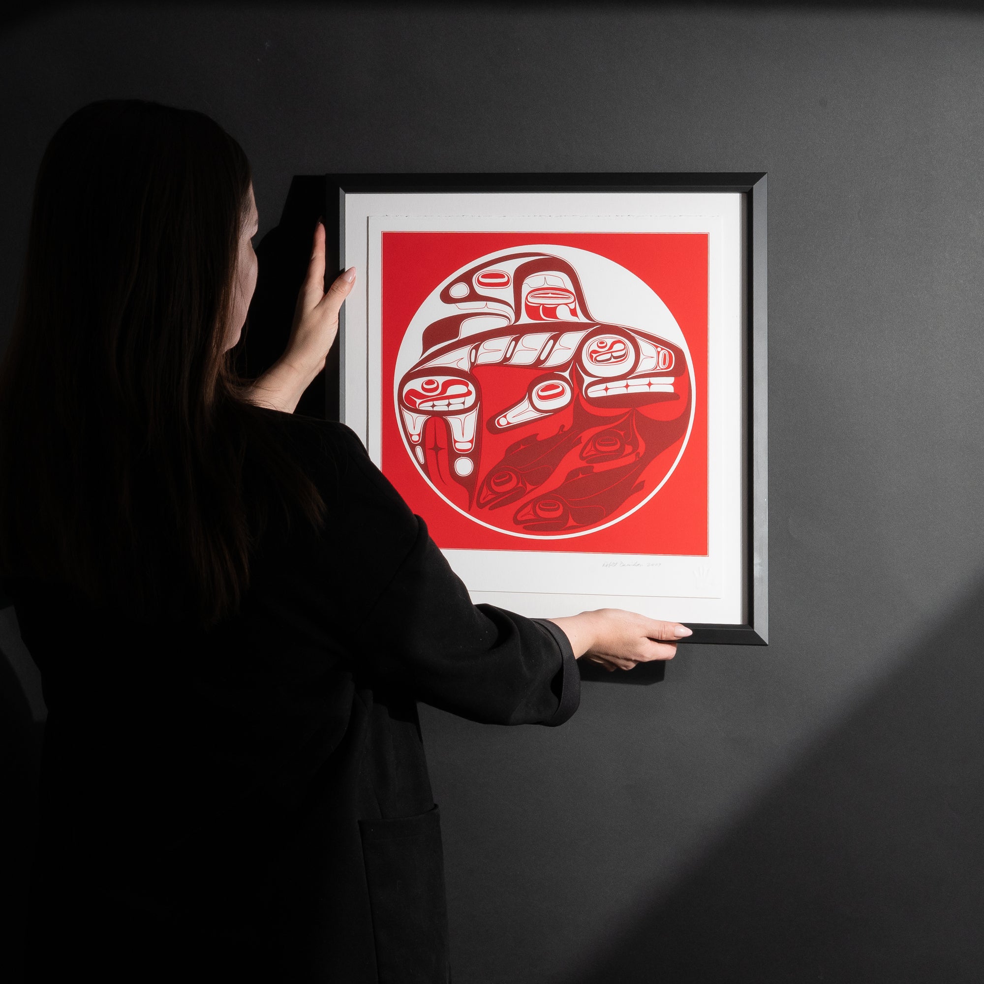 Model placing Framed Killer whale and Salmon fine Art print by renowned Haida Artist, Robert Davidson on a black wall