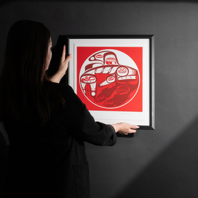 Model placing Framed Killer whale and Salmon fine Art print by renowned Haida Artist, Robert Davidson on a black wall