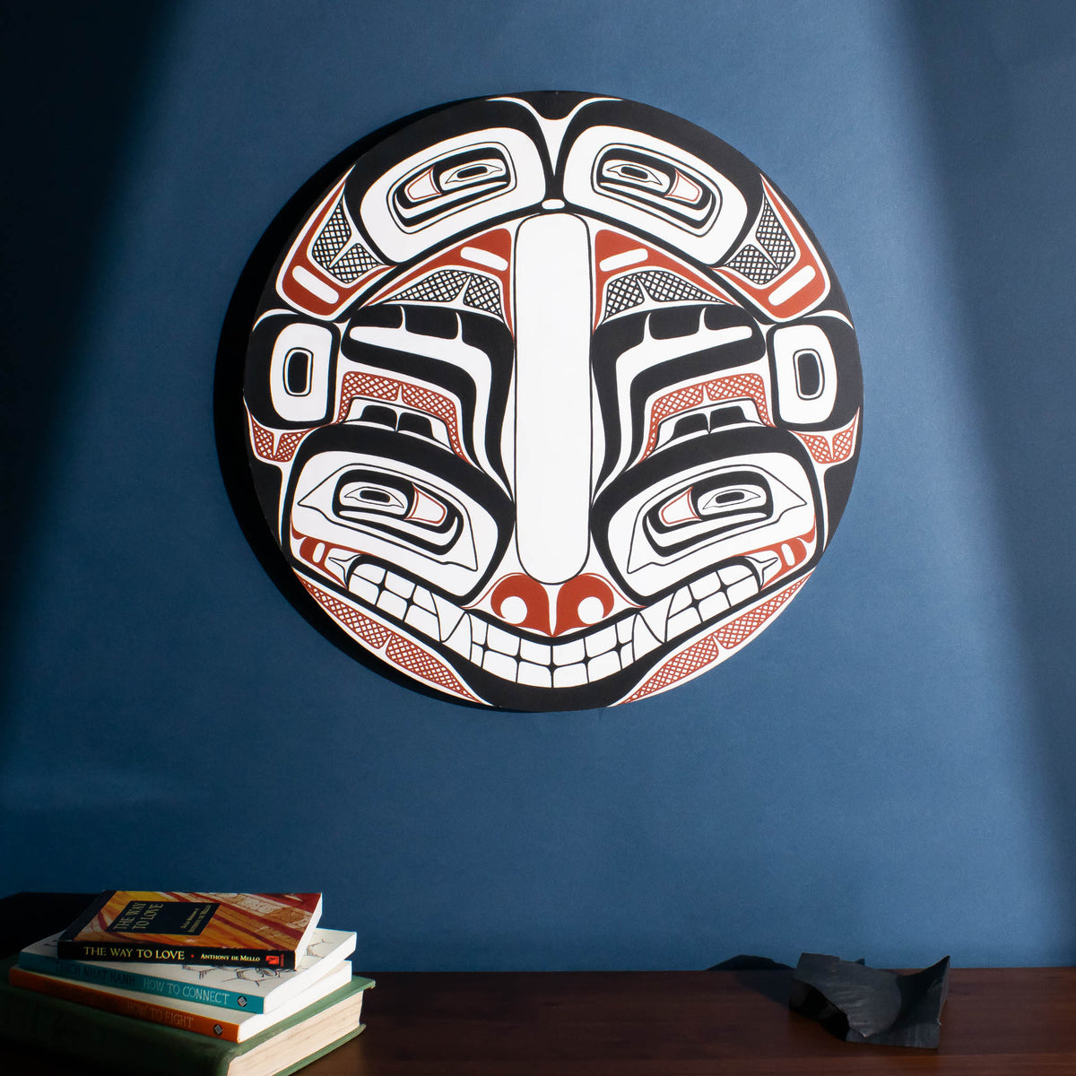 A red and black round northwest coast painting by Shoshannah Greene above a table with books and a rock.