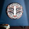A red and black round northwest coast painting by Shoshannah Greene above a table with books and a rock.