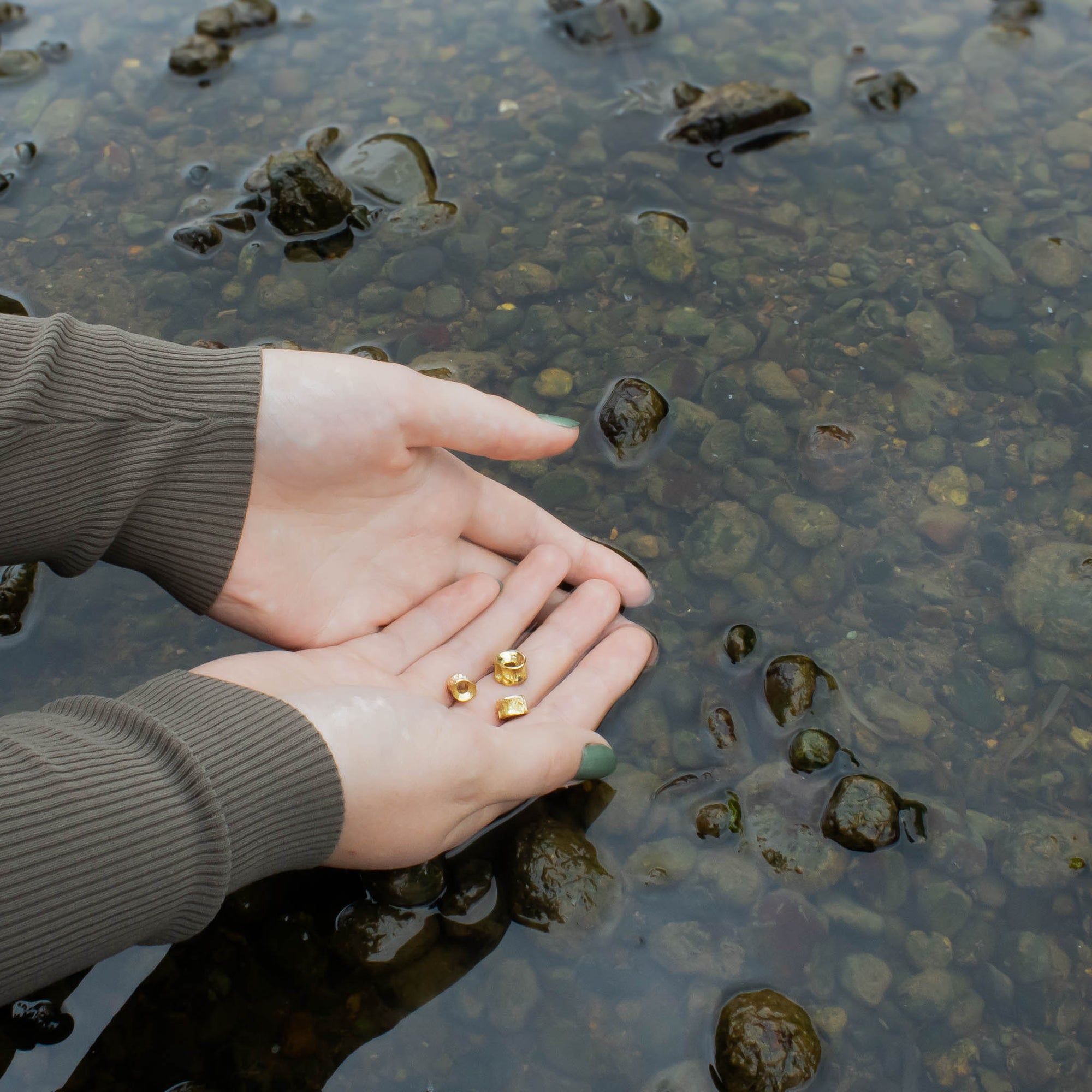 Hands holding small 10k Gold Salmon bone Vertebrae by Morgan Asoyuf over a the Tlell river with pebbles