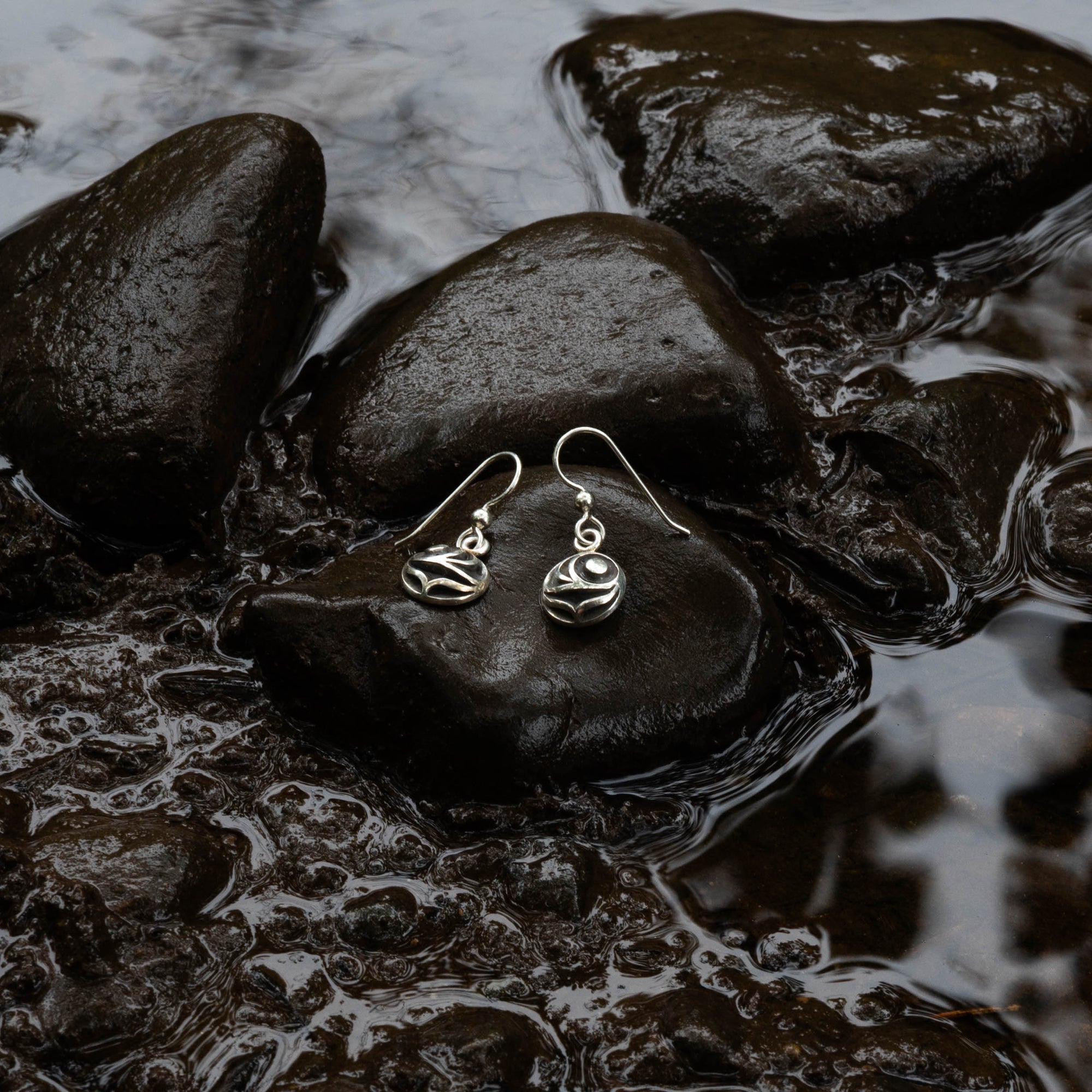 Pair of silver salmon egg earrings with black and white pattern by Indigenous Canadian Artist Meaghan Mcrae on a wet stone with water and rocks in the background