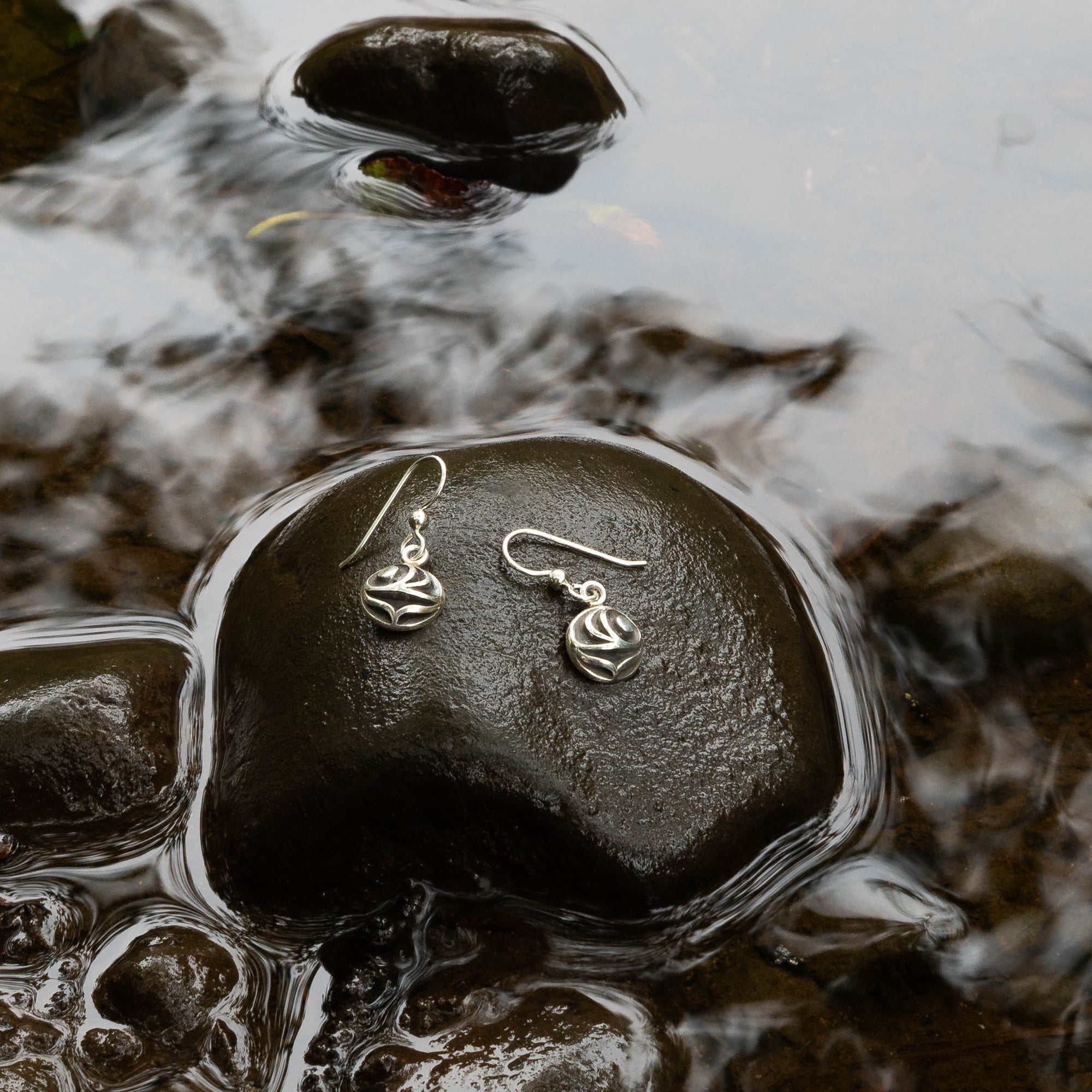Pair of silver salmon egg earrings with black and white pattern by Indigenous Canadian Artist Meaghan Mcrae on a wet stone with water and rocks in the background