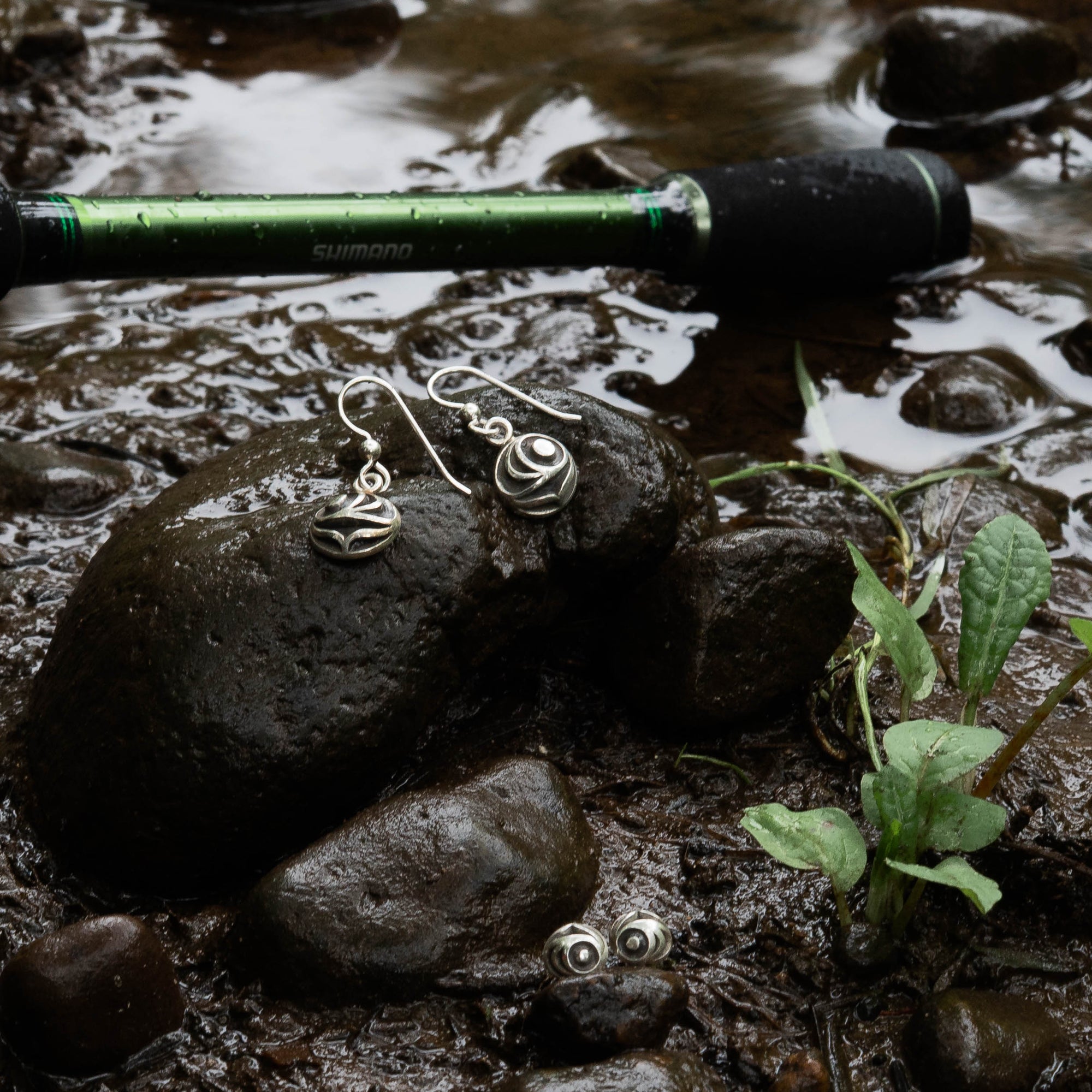 Pair of silver salmon egg earrings with black and white pattern by Indigenous Canadian Artist Meaghan Mcrae on a rock with a fishing rod in the background