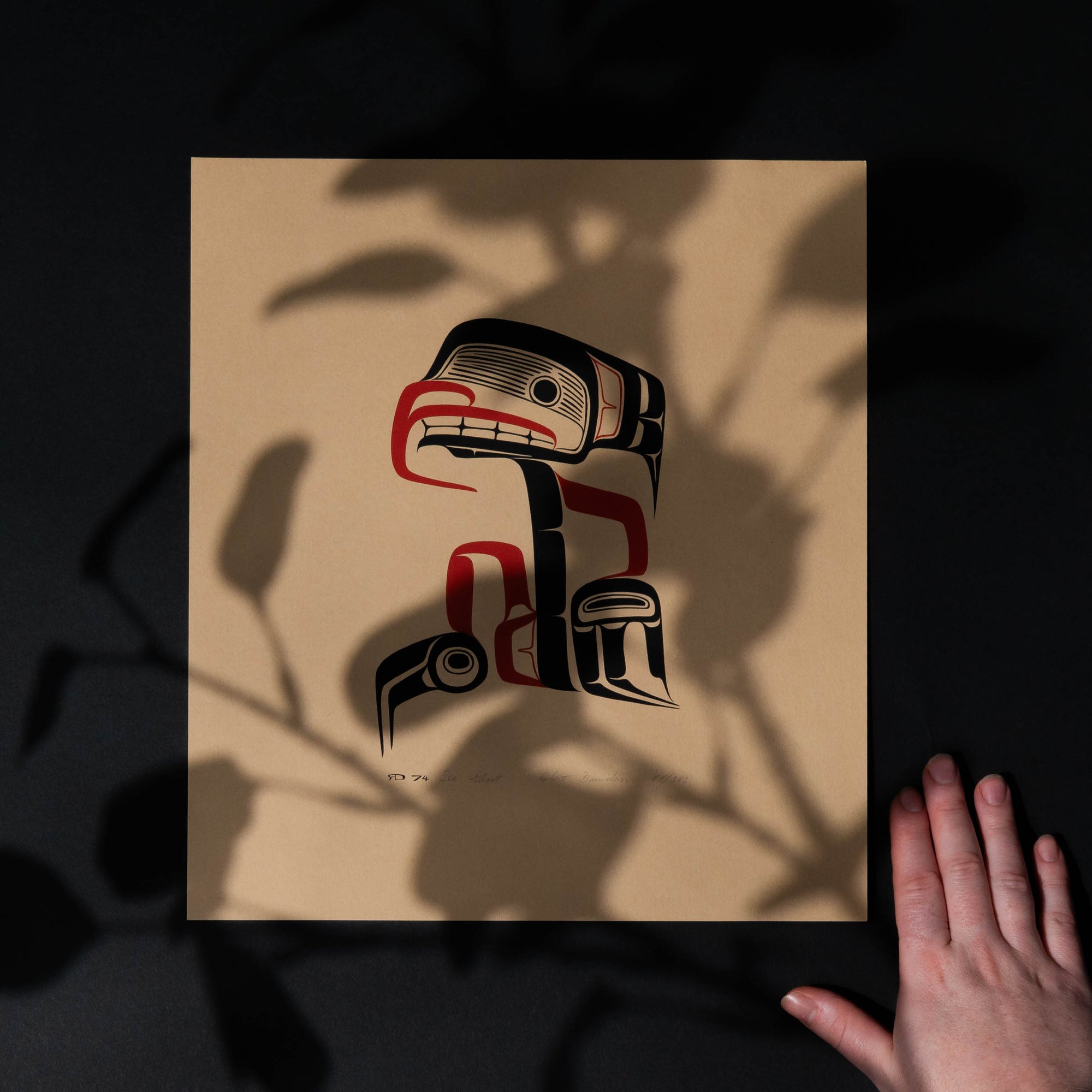 Artistic depiction of a stylized Haida Sea Ghost with red and black patterns on a beige background with a hand for scale.