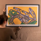Person holding a framed abstract artwork with geometric shapes on a yellow background
