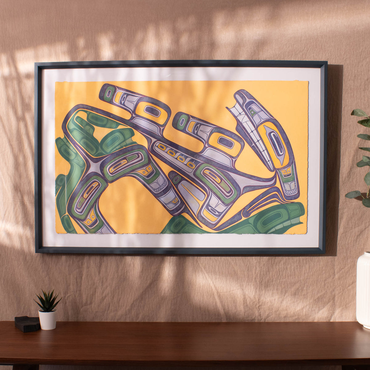 Framed abstract artwork with green and purple design on a yellow background, displayed on a wall.