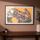 Framed abstract artwork on a wall with a wooden table and plant in the foreground.