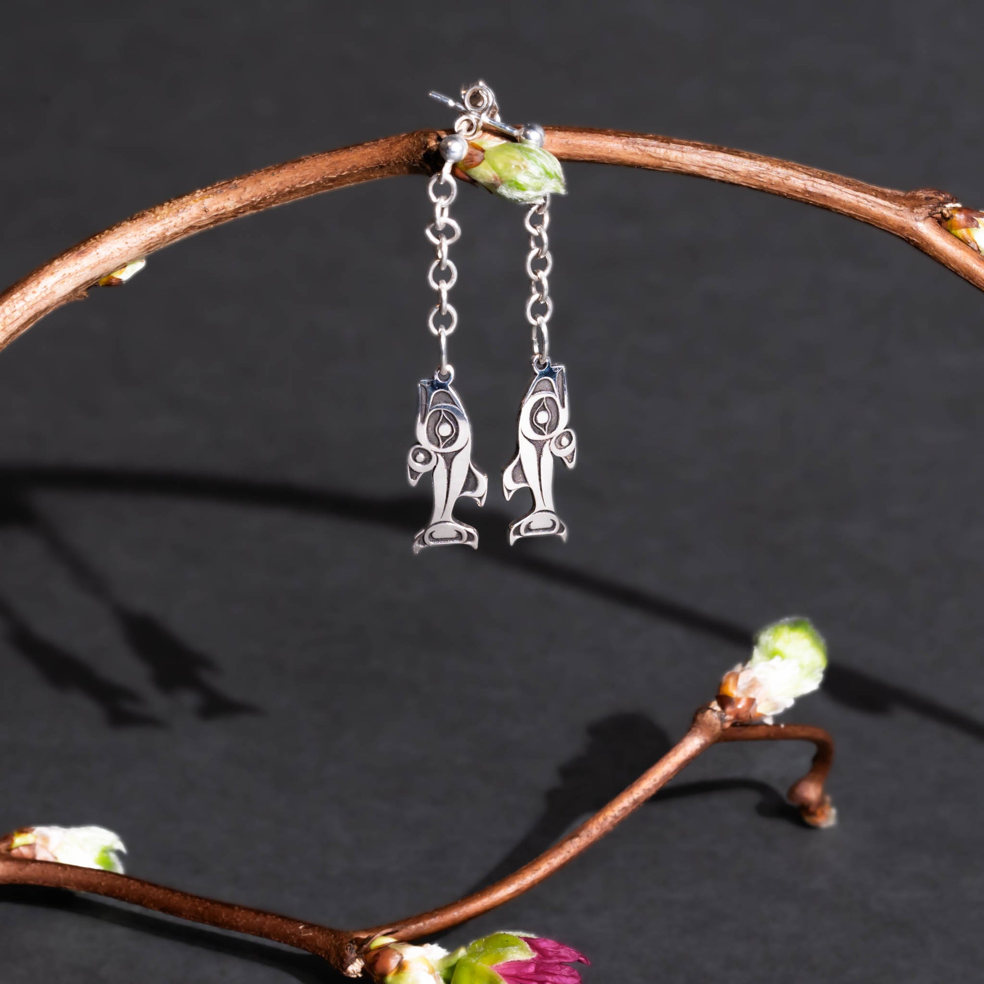 Silver salmon stud earrings with chain dangling from a twig with blossoms on it on a dark gray background.