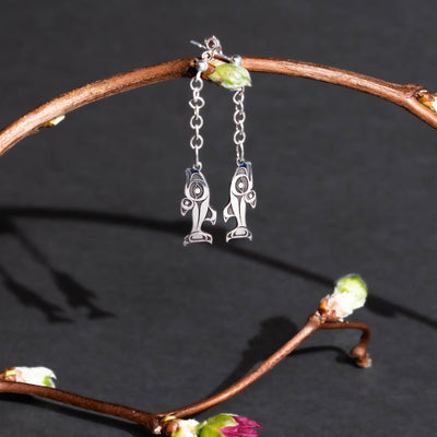 Silver salmon stud earrings with chain dangling from a twig with blossoms on it on a dark gray background.