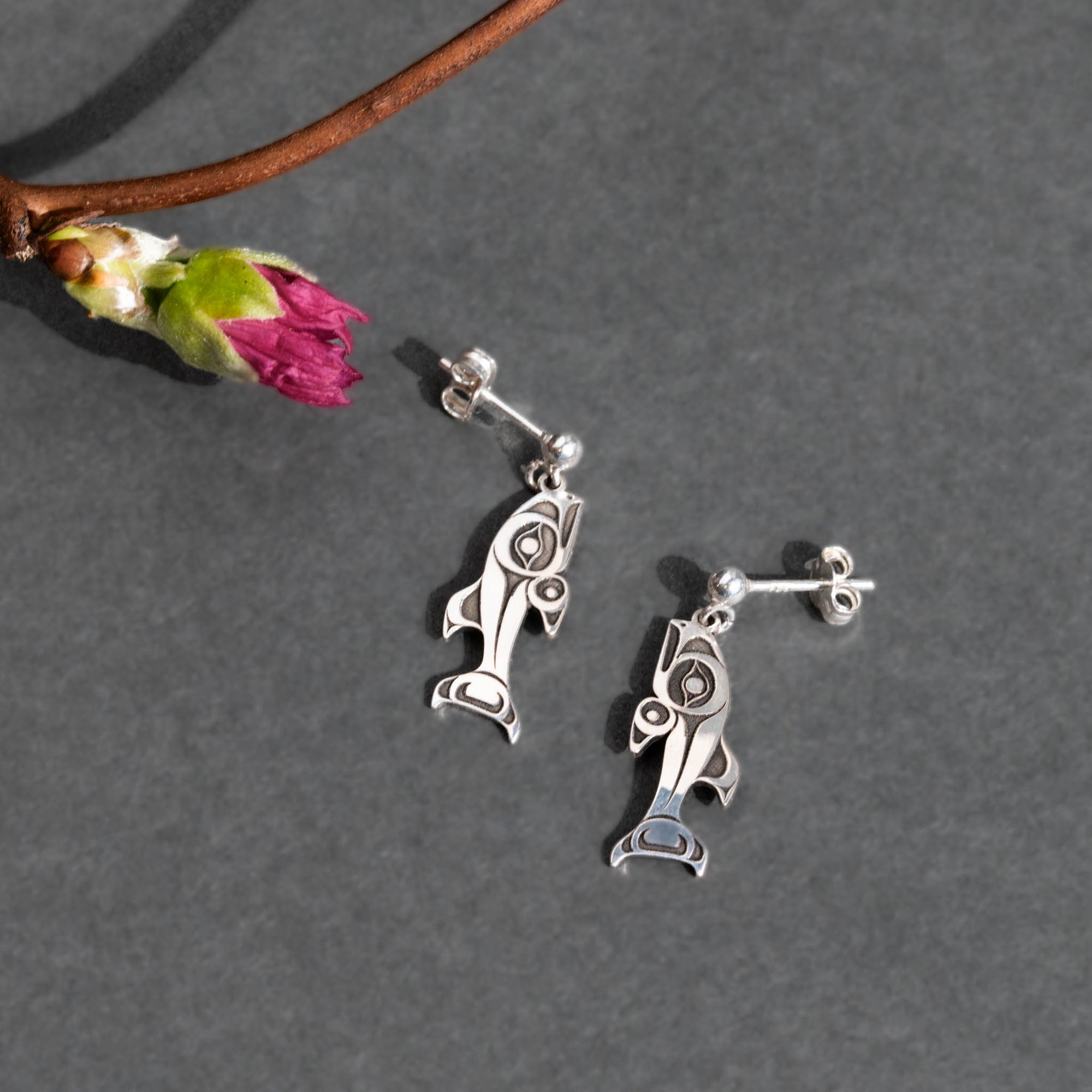 Silver fish-shaped earrings on a gray surface with a branch and flower.