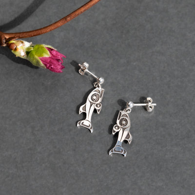 Silver fish-shaped earrings on a gray surface with a branch and flower.