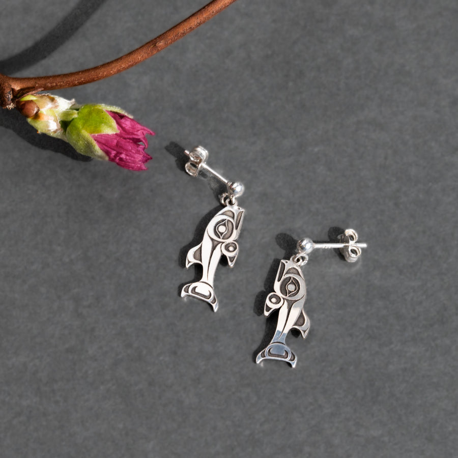 Silver fish-shaped earrings on a gray surface with a branch and flower.