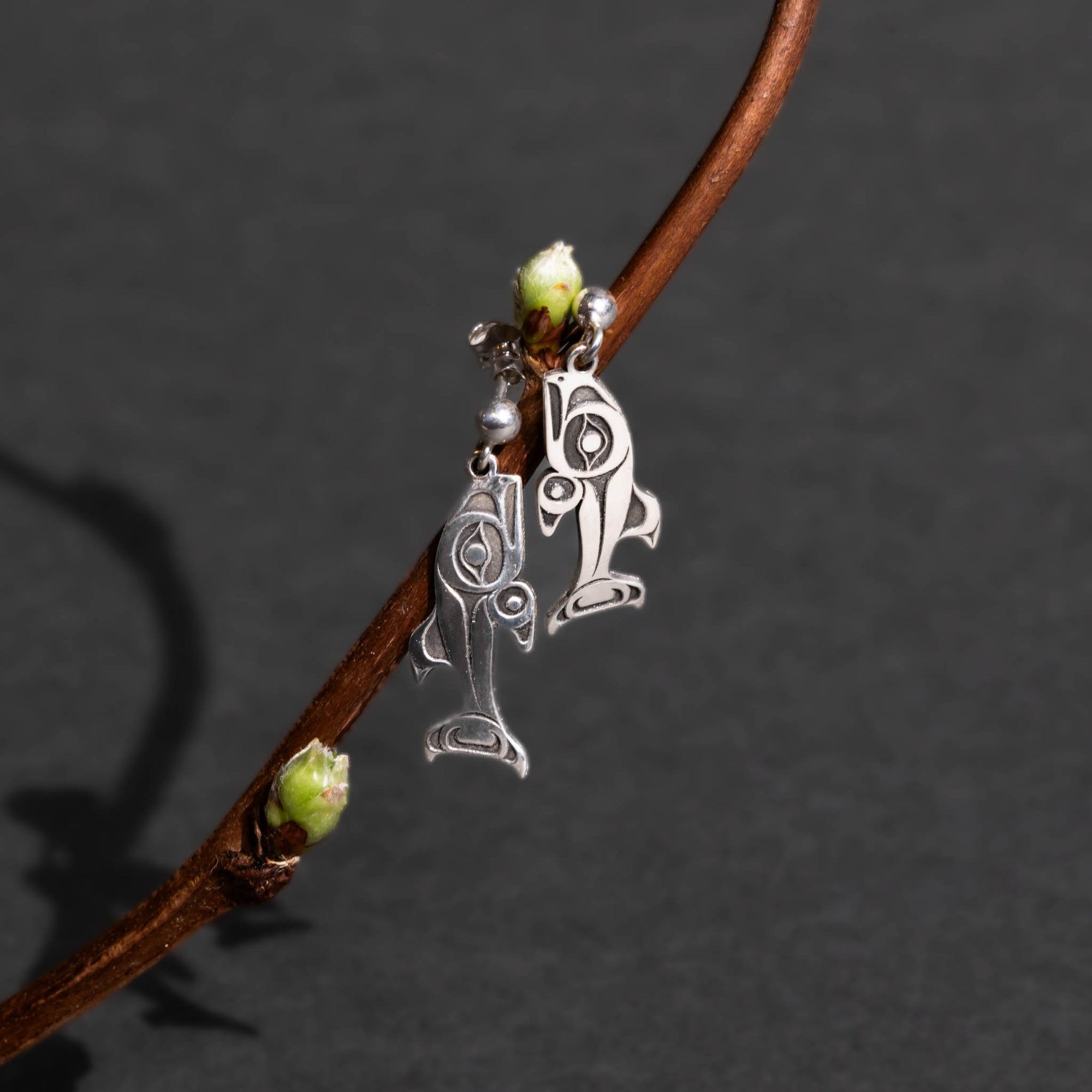 Silver salmon stud earrings dangling from a blossoms on a twig on it on a dark gray background.