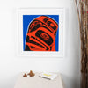 Three finned #2 in a 3 series fine art print by local Haida, Haida Gwaii Artist featuring a whale fin a human face and a raven, in blue red and black, in a white wood frame. In interior design