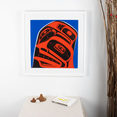 Three finned #2 in a 3 series fine art print by local Haida, Haida Gwaii Artist featuring a whale fin a human face and a raven, in blue red and black, in a white wood frame. In interior design