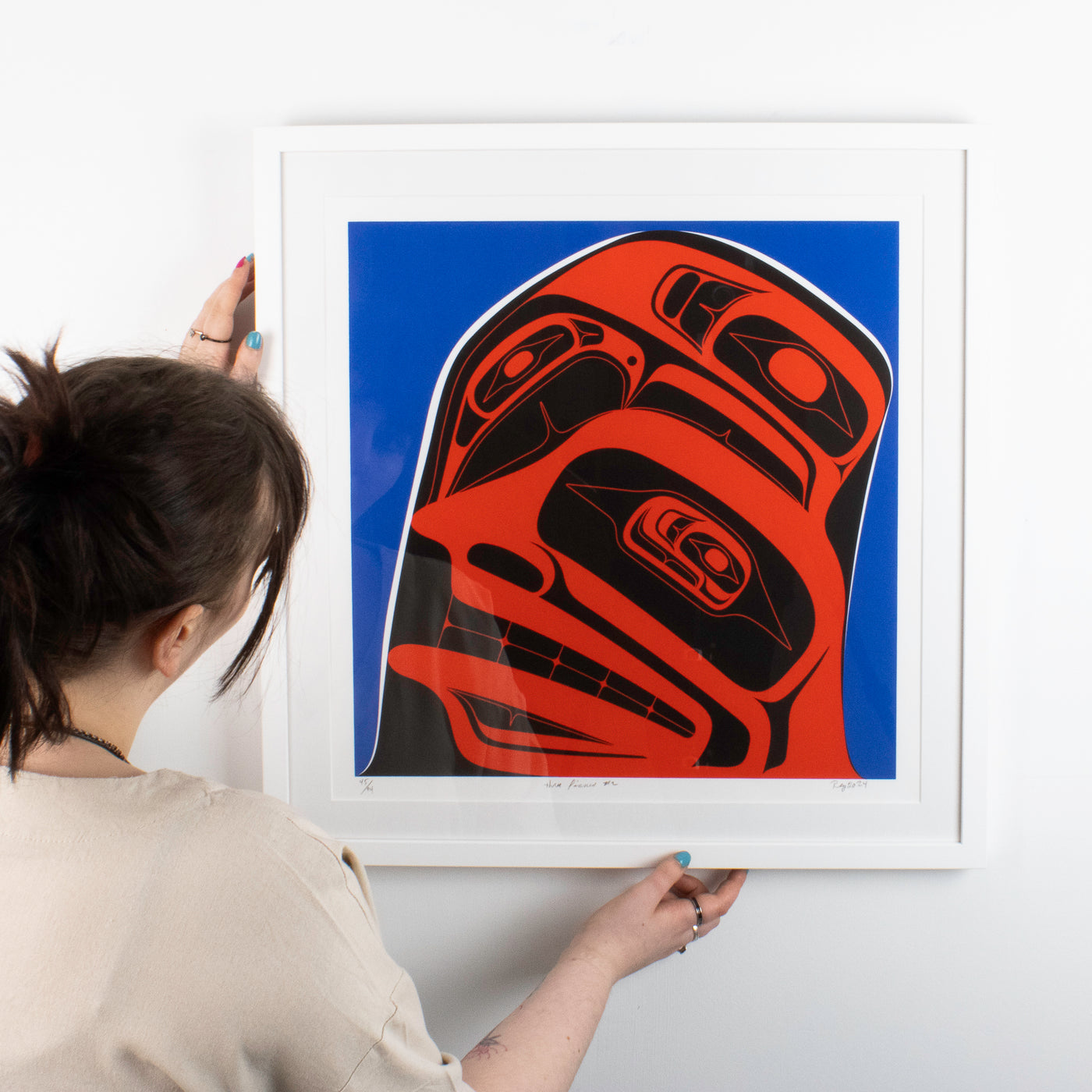 Three finned #2 in a 3 series fine art print by local Haida, Haida Gwaii Artist featuring a whale fin a human face and a raven, in blue red and black, in a white wood frame. With a person for size ratio