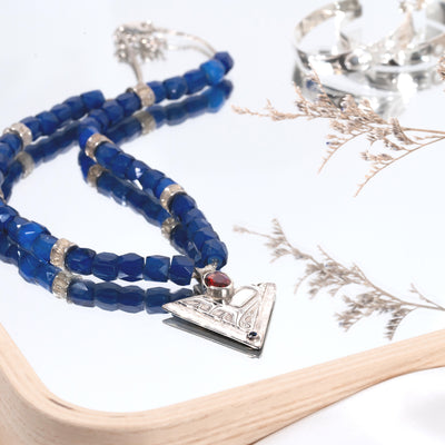 Russian Blue trade beads and Salmon bone vertebrae beaded necklace with silver clasp and silver triangular pendant featuring a formline hummingbird, with garnet and sapphire on a white surface with dried plants.