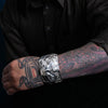 Person wearing a silver bracelet northwest coast bracelet with tattoos on their arm against a black background
