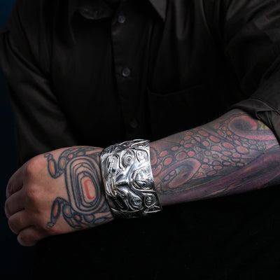 Person wearing a silver bracelet northwest coast bracelet with tattoos on their arm against a black background