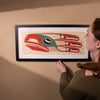 Woman looking at a framed artwork of a Framed fine artwork of a stylized bird with red, green, and black colors on a beige background by Haida Artist Robert Davidson