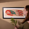 Person looking at a Framed fine artwork of a stylized bird with red, green, and black colors on a beige background by Haida Artist Robert Davidson