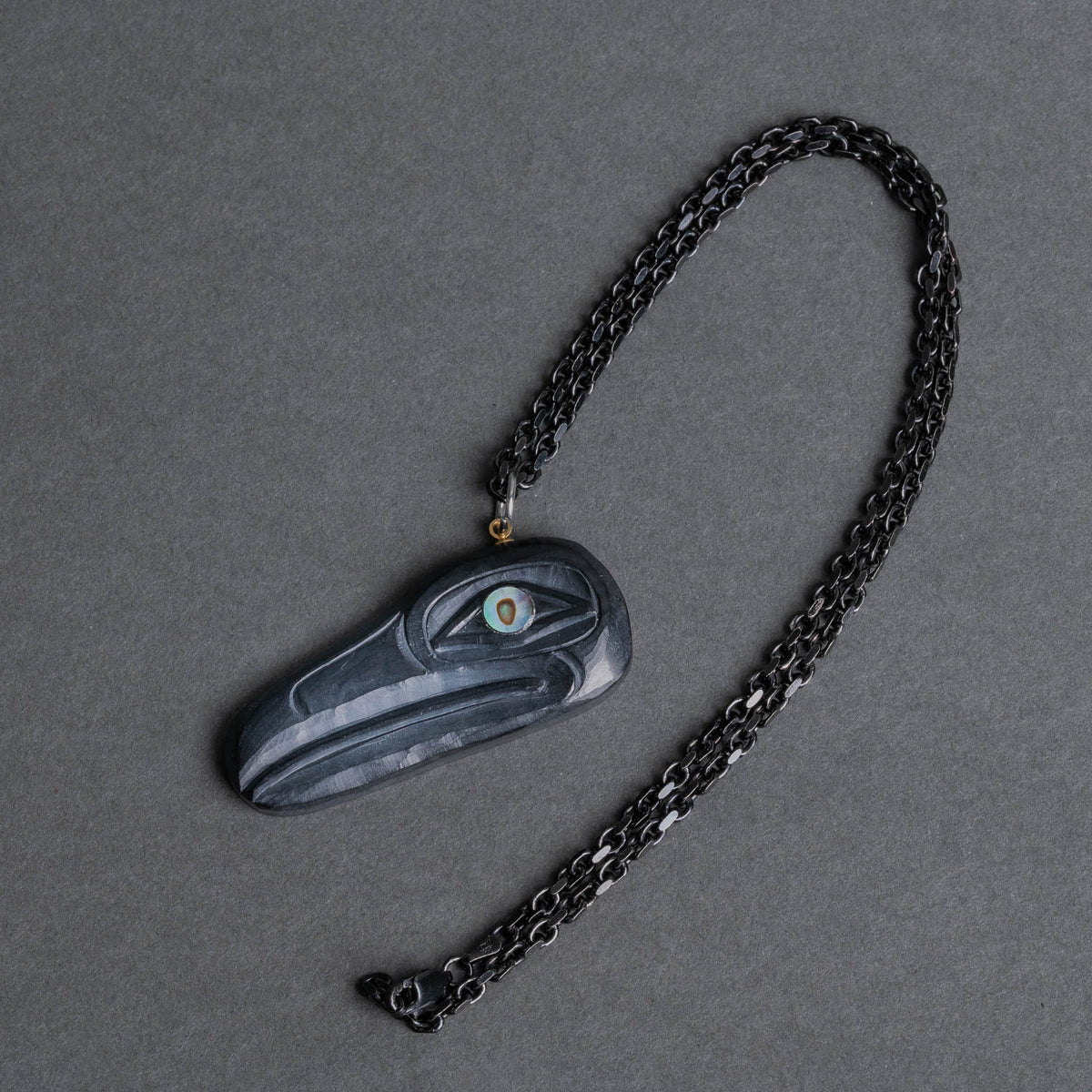 A dark charcoal gray carved raven pendant  with a black chain, displayed on a textured gray background.