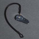 A dark carved raven pendant with a small iridescent eye detail, on a black chain on a textured gray background.