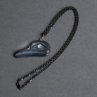 A dark carved flicker necklace on a dark metal chain, displayed on a textured gray background.