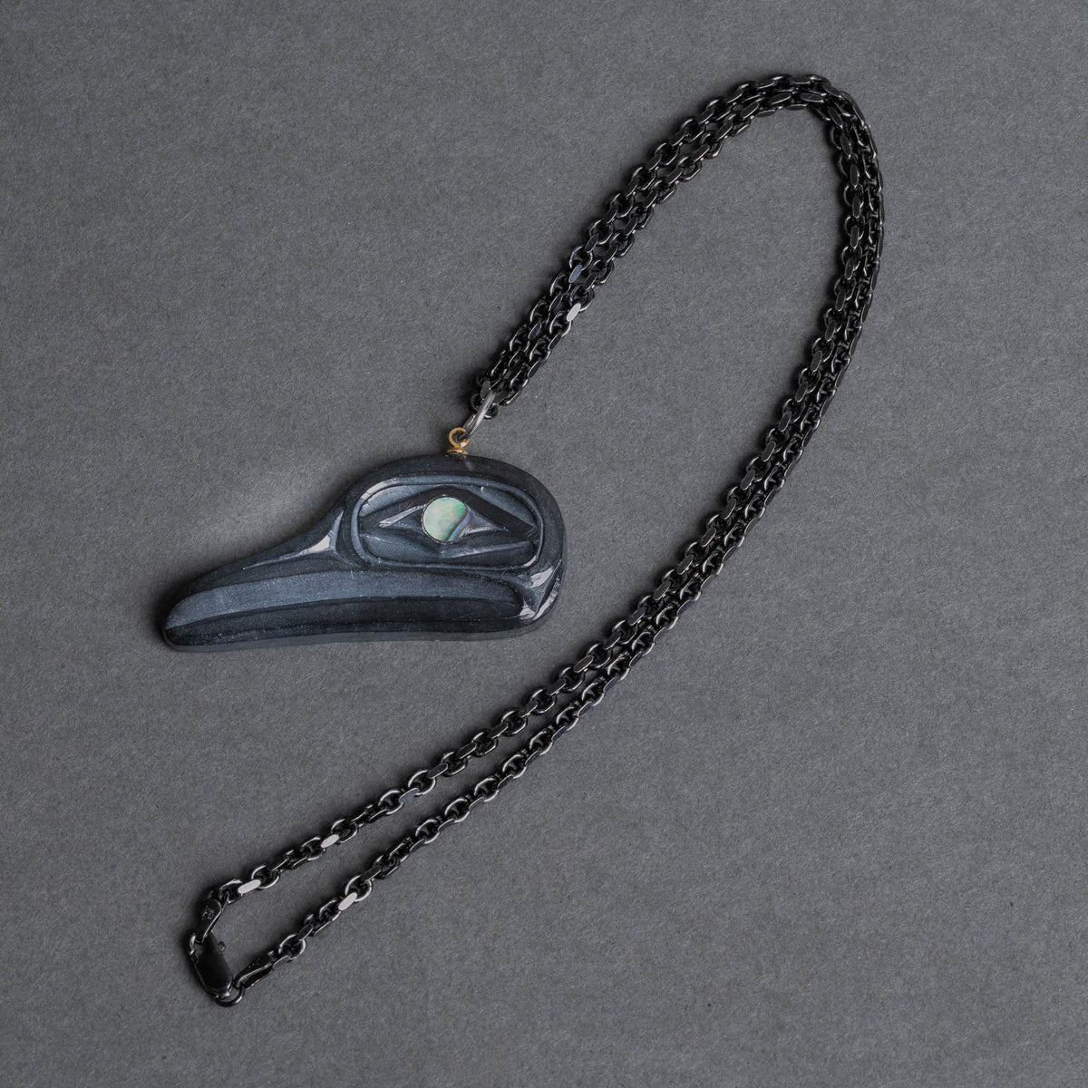 A dark carved flicker necklace on a dark metal chain, displayed on a textured gray background.