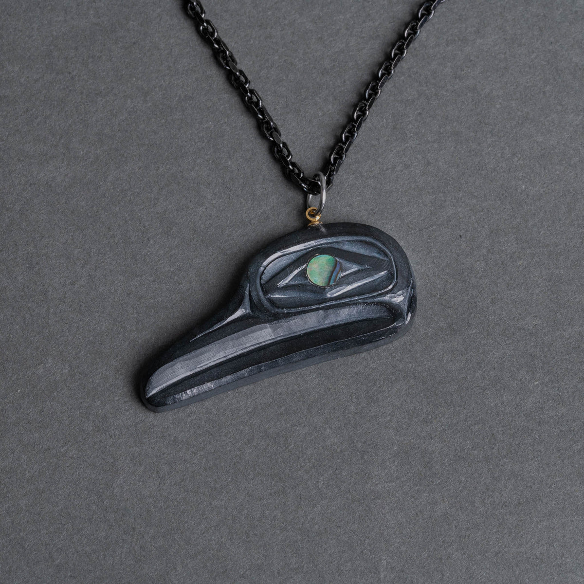 A black flicker pendant necklace with a small iridescent green eye detail, shown on a dark gray textured background.
