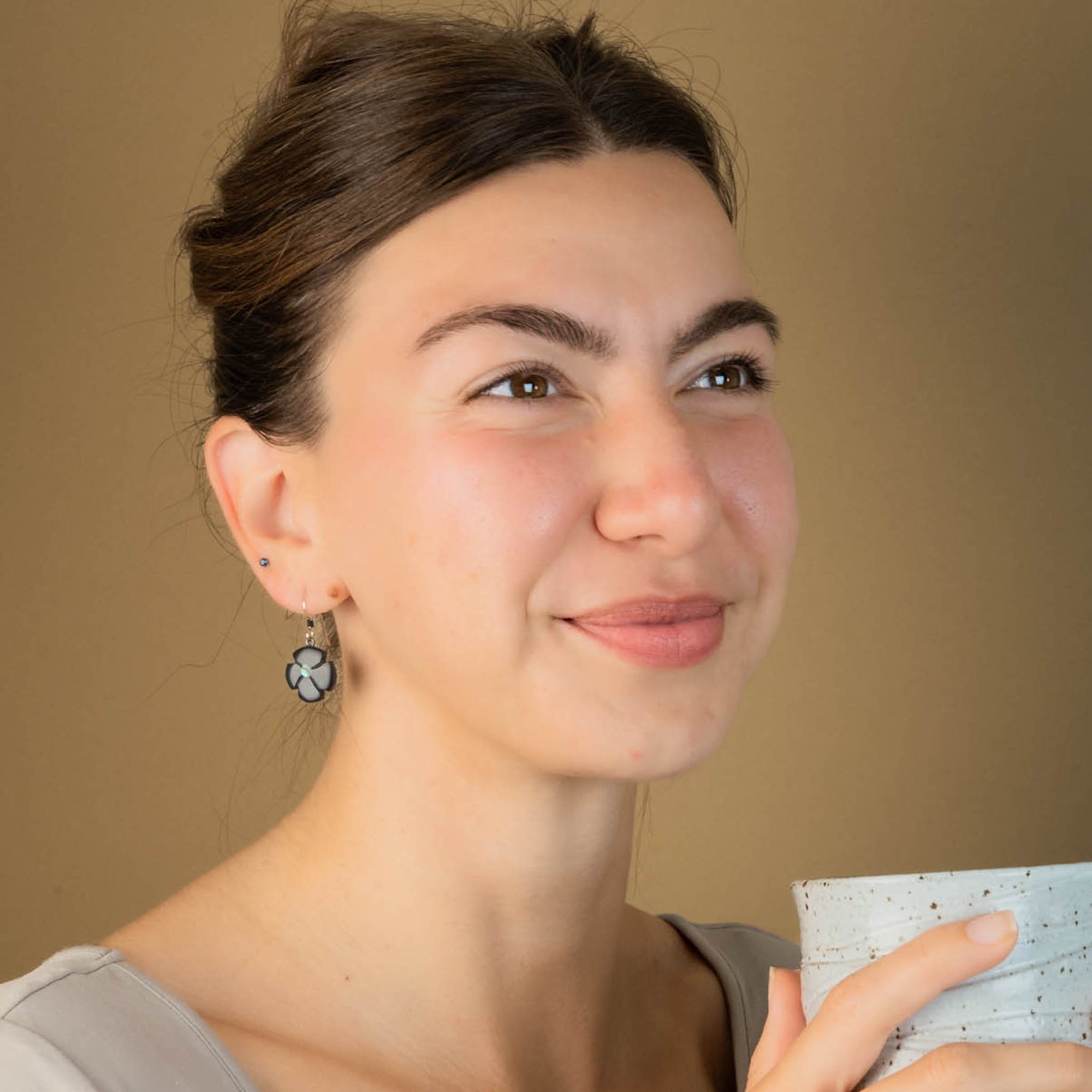 Woman holding a coffee cup against a beige background facing right and wearing black flower shaped earrings.