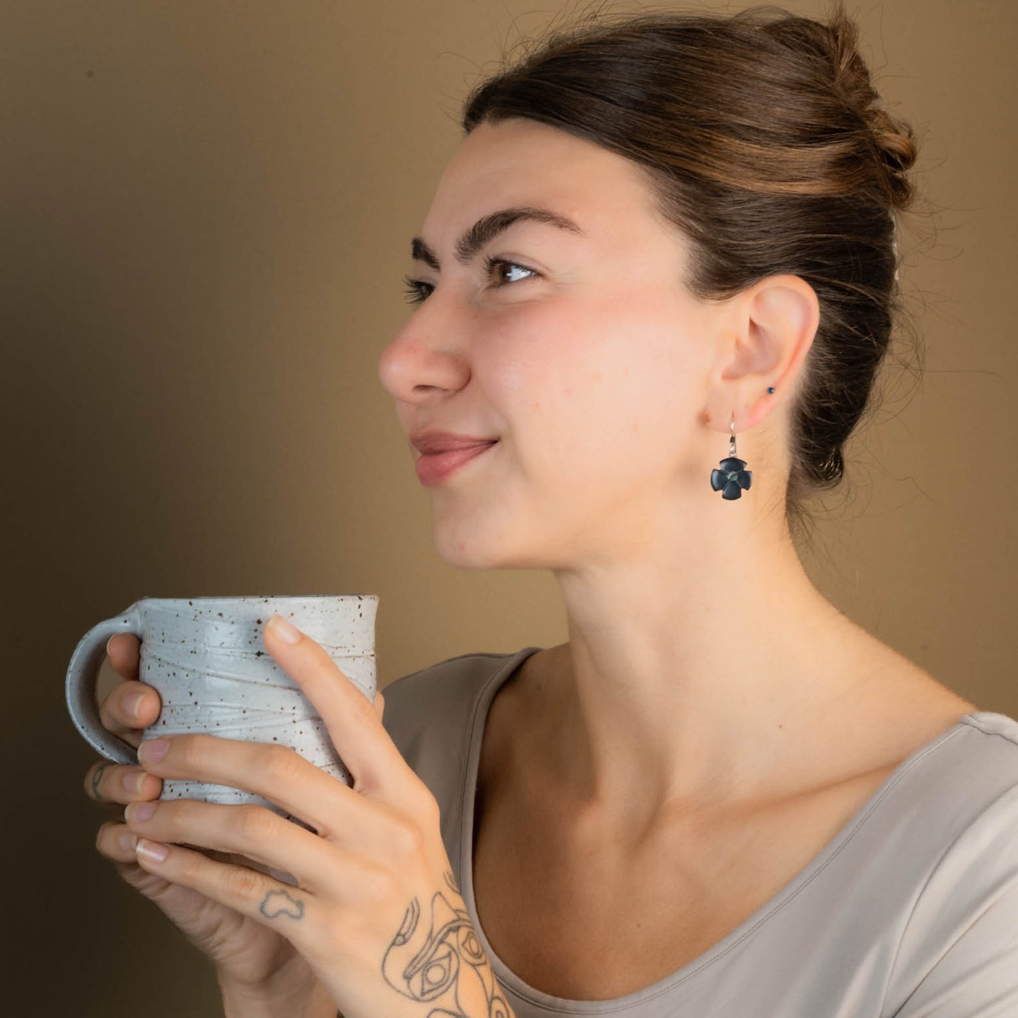 Woman facing left holding a textured mug against a plain background wearing a grey shirt and black flower earrings.