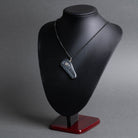 A dark gray stone pendant with a gold accent, displayed on a matte black jewelry bust with a dark wood base against a gray background.