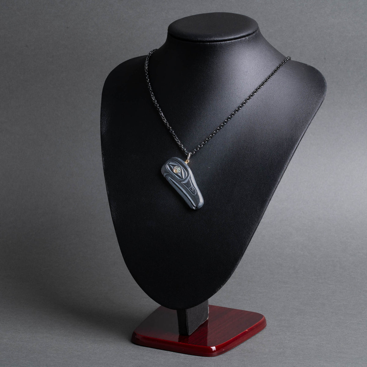 A dark gray stone pendant with a gold accent, displayed on a matte black jewelry bust with a dark wood base against a gray background.