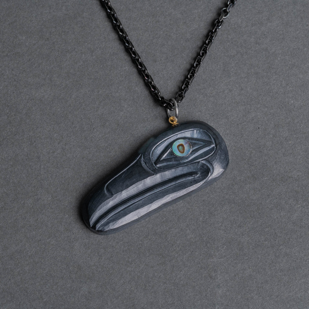 Carved black raven pendant with shimmering eye and black chain on a dark background facing to the right.