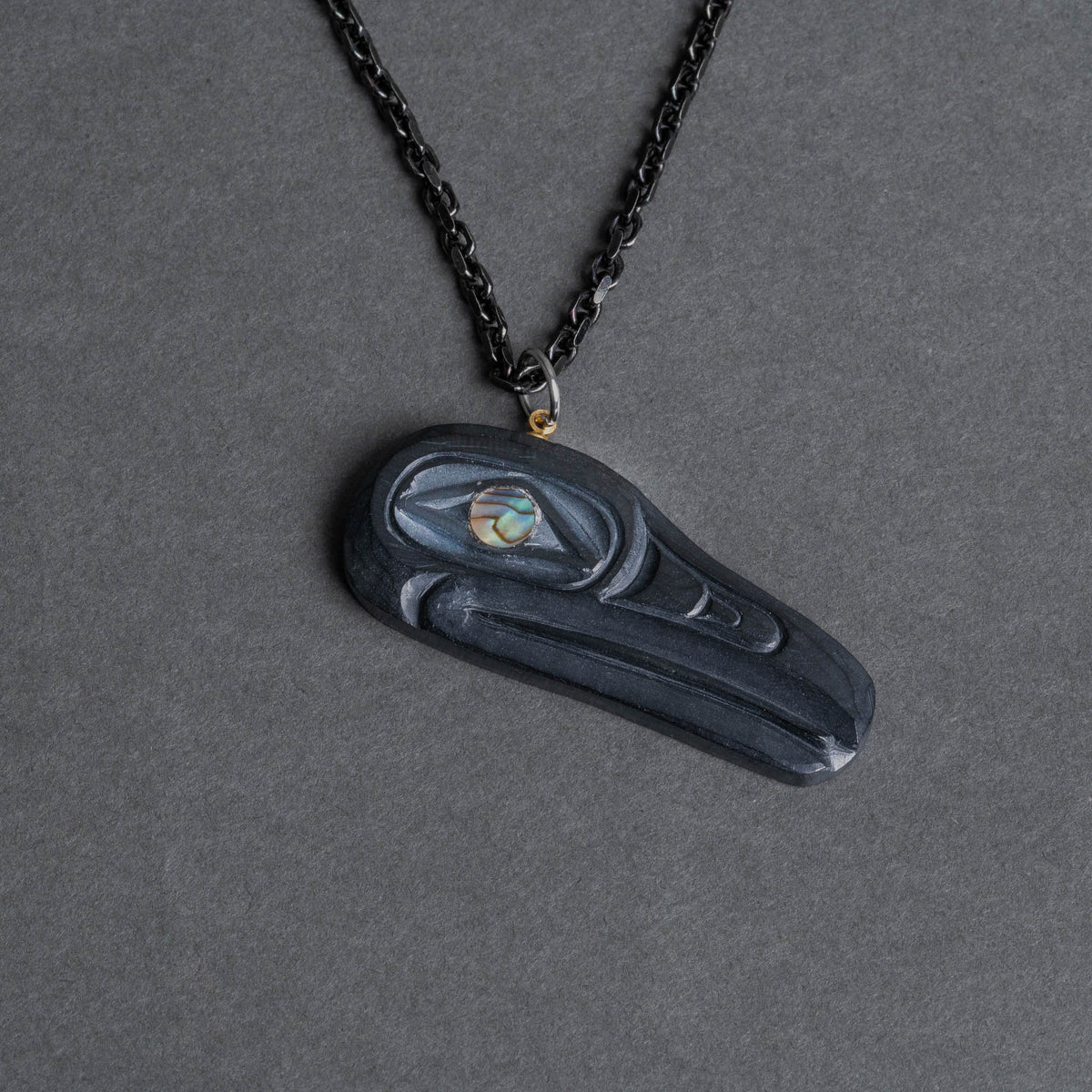 Carved black raven pendant with shimmering eye and black chain on a dark background.