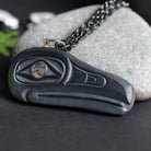 close up of a carved stone featuring a black raven, and multi coloured eye, with a black chain set against a light grey rock on a black and green  back ground.