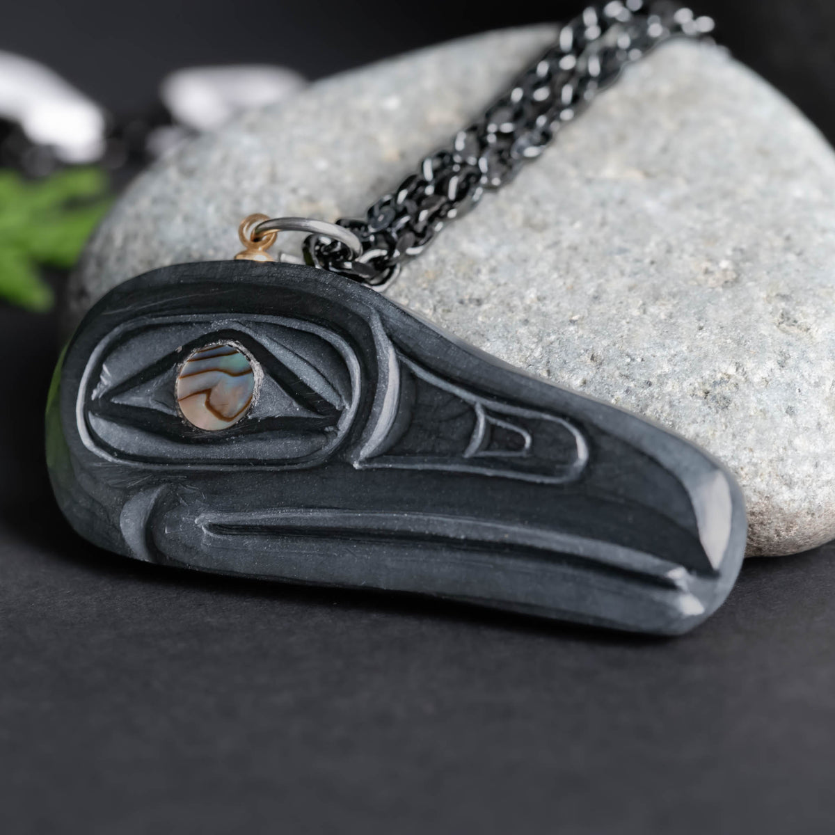 close up of a carved stone featuring a black raven, and multi coloured eye, with a black chain set against a light grey rock on a black and green  back ground.