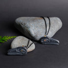 Two dark grey stone necklaces proped against light grey rocks with a cedar sprig beside them, one a dork grey backgound.