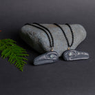 Raven pendants facing oppisite ways with an irradecent eye set against a grey rock with a cedar sprig on  dark grey background.